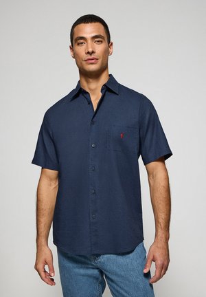 Camisa de mangas cortas en color marino, fabricada en un tejido ligero. Cuenta con un cierre frontal de botones y un bolsillo en el pecho con un pequeño detalle de logo en rojo. Textura suave.