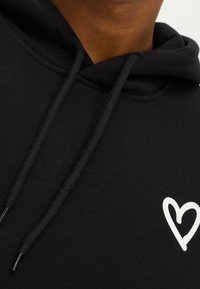 Felpa nera in cotone con un grafico a forma di cuore bianco, scollatura rotonda e lacci con estremità testurizzate. Tessuto morbido, stile informale.