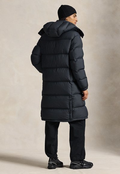 Μαύρο παλτό puffer με κουκούλα, τετράγωνο σχήμα και ραμμένη υφή. Φορεμένο με μαύρο παντελόνι και αθλητικά παπούτσια που ταιριάζουν. Ουδέτερο φόντο.