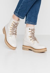 Witte glanzende leren enkellaarsjes met een nepbont kraag, decoratieve gouden veterogen en bruine veters op een gestructureerde rubberen zool.