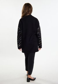 Cardigan lungo nero con lavorazione a maglia testurizzata, caratterizzato da accenti di filo metallico con motivi a onda sulle maniche. Combinato con pantaloni neri aderenti e tacchi.