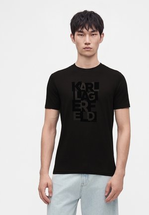 Ung mand med kort sort hår iført sort Karl Lagerfeld T-shirt og lyseblå jeans mod en ensfarvet hvid baggrund.