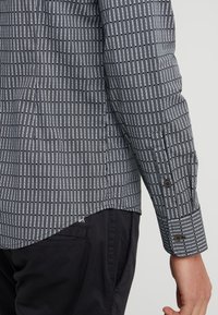 Camicia a maniche lunghe realizzata in tessuto a stampa bianco e nero con strisce verticali e testo. Dettaglio dei bottoni neri sul polsino.