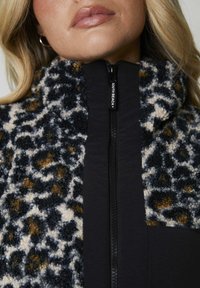 Fleecejacka med en texturerad leopardmönster i beige, svart och brun, med en svart dragkedja och hög krage.