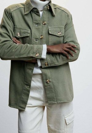 Personne à la peau foncée portant une veste verte boutonnée par-dessus un pull à col roulé blanc et un pantalon de couleur claire avec une poche latérale.