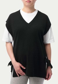 Svart stickad ärmlös tröja med V-ringning, sidofästen och ribbade kanter. Lagd över en vit T-shirt och kombinerad med grå byxor.