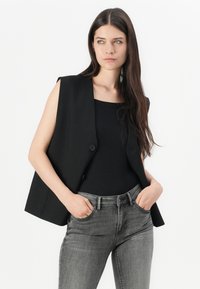 Black sleeveless blazer with a single button, smooth fabric, paired with a black top and gray fitted jeans, highlighting a streamlined silhouette.