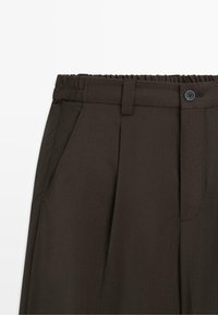 Dark brown pants with elastic waistband, belt loops, front side pocket, button closure, and front pleats visible on white background.
