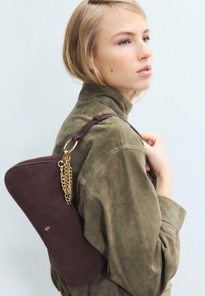 Femme blonde portant une veste vert olive et tenant un sac à bandoulière marron foncé avec des détails en chaîne dorée, sur un fond clair uni.