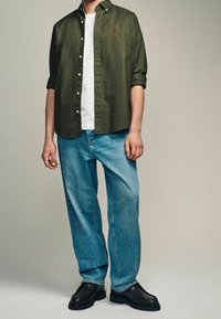 Uomo che indossa una camicia verde oliva con bottoni sopra una maglietta bianca, jeans azzurri chiari e scarpe nere, in piedi contro uno sfondo neutro.