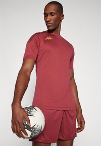 Bordeaux sportshirt met korte mouwen, gouden accenten op de schouders en bijpassende korte broek. Model houdt een zwart-witte voetbal vast.