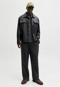 Chaqueta de cuero negra sobre una camisa de cuadros, pantalones negros holgados, zapatos marrones y una gorra verde claro, que lleva puesta una persona de pie frente a un fondo blanco.