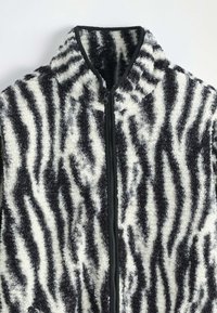 Veste en polaire zebra rayée noir et blanc avec un col montant et une fermeture éclair à l'avant, présentant une surface texturée et pelucheuse.