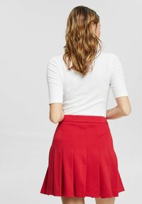 Jupe plissée rouge avec une taille ajustée et une texture douce, accompagnée d'un haut à manches courtes blanc. La vue de dos met en valeur la forme de la jupe.