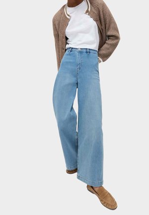 Osoba w jasnoniebieskich dżinsach z wysokim stanem i szerokimi nogawkami, białej koszulce, brązowym kardiganie oraz brązowych mokasynach.