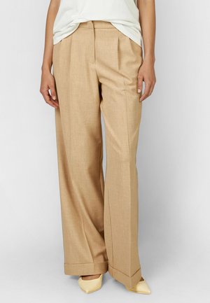 Woman wearing beige wide-leg trousers with front pleats, paired with a white top and cream pointed-toe heels against a plain background.