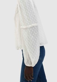 Blusa bianca leggera con design a pois testurizzati, maniche lunghe svasate e polsini arricciati, abbinata a jeans blu scuro.