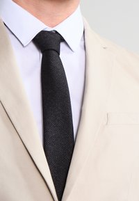 Chaqueta de traje beig con un único bolsillo, camisa blanca y una corbata negra texturizada. Enfocado en líneas limpias y un ajuste a medida sin patrones.
