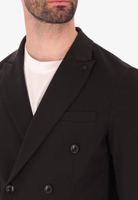Blazer nero doppiopetto con rever a punta, dettagli con bottoni e una tasca; indossato sopra una maglietta bianca a collo rotondo. Tessuto dalla superficie liscia.