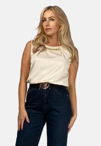 Kremowy top bez rękawów z akcentem w postaci złotego łańcuszka, zestawiony z ciemnoniebieskimi spodniami o wysokim stanie oraz czarnym paskiem ze złotą klamrą.