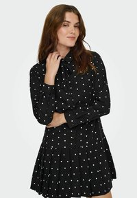 Juoda suknelė su baltais taškeliais, su apykakle, sagomis priekyje ir klostuota sijono konstrukcija. Pagaminta iš lengvos medžiagos.