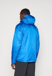 Modra izolirana jakna s kapuco, s gladko, sijočo teksturo in podaljšanimi rokavi, zasnovana za zunanjo uporabo.