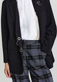 Blazer nero con dettagli in metallo argentato e taschino, abbinato a pantaloni a quadri grigi con cintura e tasche. Camicia bianca con pieghe sotto.