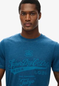 Camiseta de algodón azul con cuello redondo, que presenta texto impreso en azul más oscuro. La camiseta tiene mangas cortas y una textura suave.