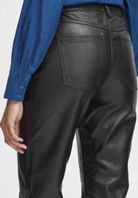 Zwarte leren broeken met een gladde textuur, uitgerust met twee achterzakken en zichtbare stiksel details, gecombineerd met een blauw shirt met lange mouwen.