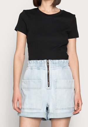 Kvinde iført en sort top med korte ærmer og højtaljede lyse denimshorts med lynlås foran og store lappe-lommer, stående med armene afslappet ned langs siden.