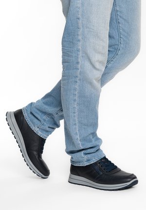 Gambe con jeans azzurri chiari e sneakers in pelle nera con suole grigie, un piede leggermente sollevato su sfondo bianco.