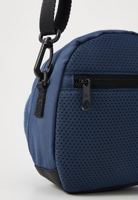 Borsa a spalla rotonda in blu navy con superficie testurizzata. Presenta una tasca laterale con zip, una tracolla regolabile e finiture in metallo nero.