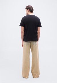 Άνδρας με την πλάτη γυρισμένη, φοράει μαύρο κοντομάνικο πουκάμισο και μπεζ παντελόνι με φαρδιές παντελόνες, στέκεται μπροστά σε απλό λευκό φόντο.