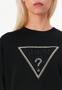 Crna dukserica s trokutastim dizajnom i teksturiranim logotipom "GUESS", ukrašena rhinestones kamenčićima i istaknutim simbolom upitnika.