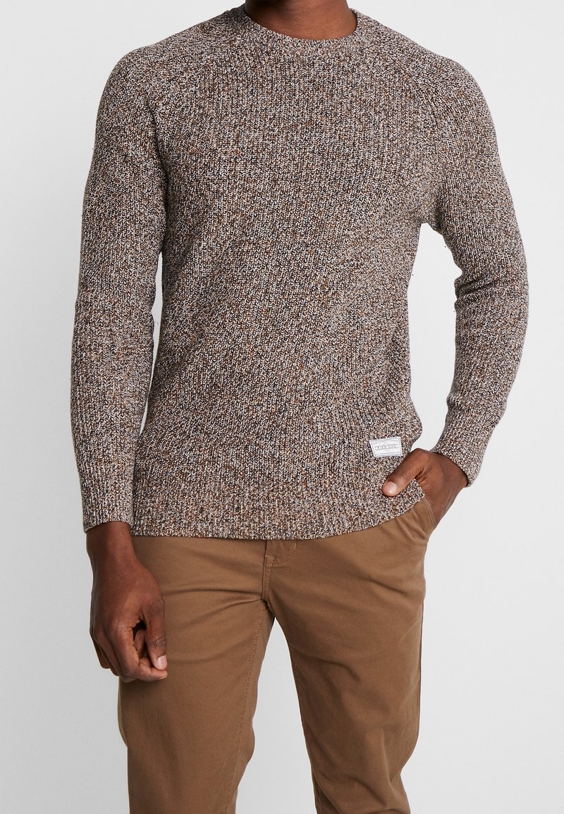 Brauner gestrickter Pullover aus strukturiertem Stoff, Rundhalsausschnitt und gerippten Bündchen. Etikett auf der unteren Kante angenäht. Kombiniert mit beigen Hosen.