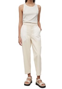 Femme portant un haut sans manches écru, un pantalon beige bicolore taille haute et des sandales compensées beiges, debout avec une main dans la poche.