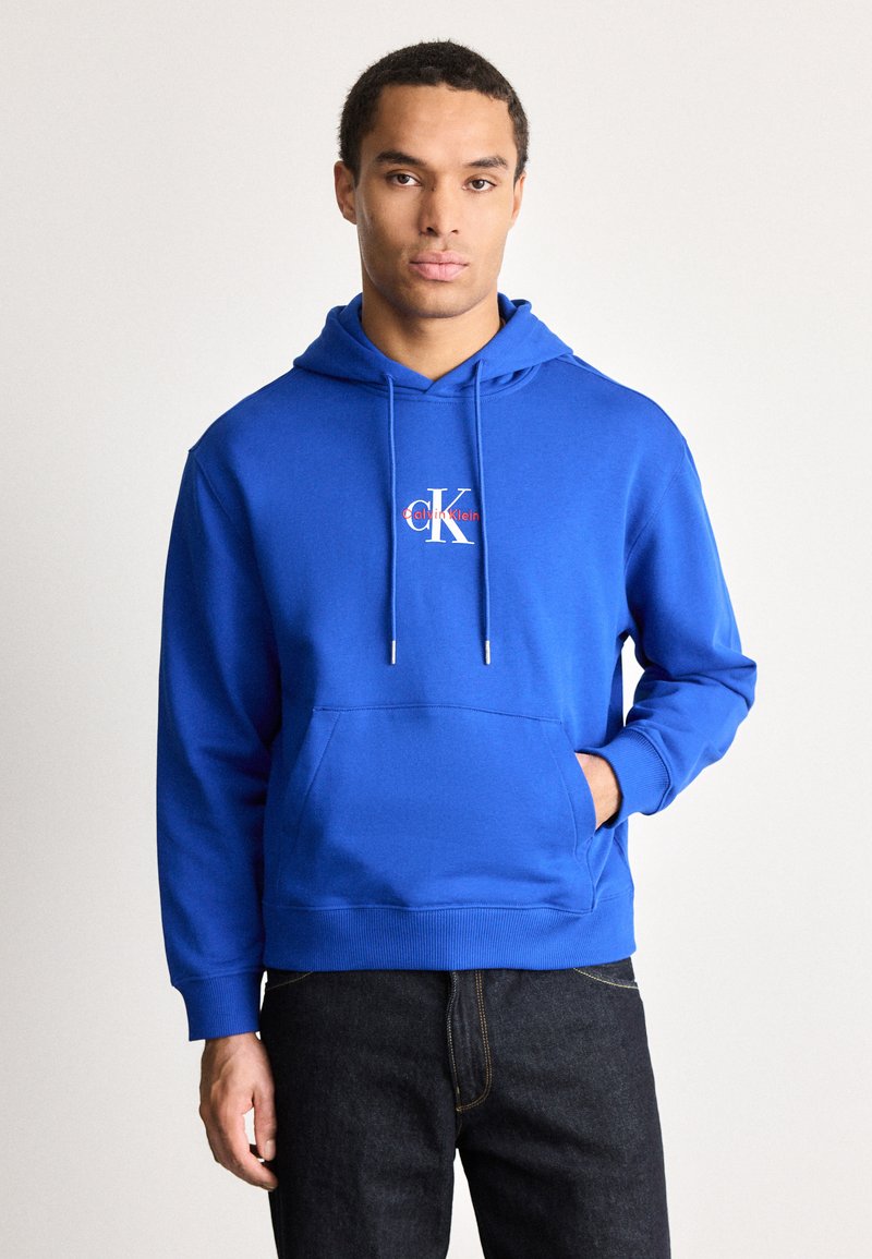 Egy férfi élénk kék Calvin Klein kapucnis pulóvert visel, elülső zsebével és sötét farmernadrággal, egy sima világos háttér előtt áll.