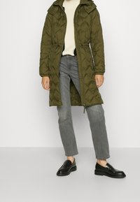 Manteau long matelassé vert olive avec fermeture éclair à l'avant, taille élastiquée et capuche. Associé à un jean gris et des mocassins noirs.