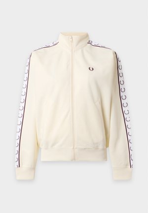 Femme debout les mains dans les poches, portant une veste crème avec des logos sur les manches, une chemise blanche, un pantalon beige large et des chaussures noires.