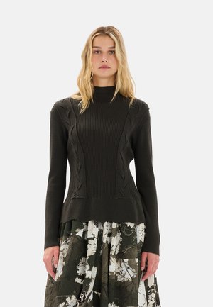 Maglione a coste di un verde scuro con collo alto e dettagli a trecce, caratterizzato da un orlo svasato. Indossato sopra una gonna con stampa floreale.