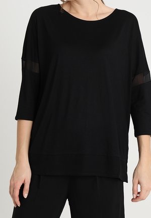 Femme portant un haut noir ample à manches trois-quarts avec des bandes horizontales transparentes près des coudes, associé à un pantalon noir.