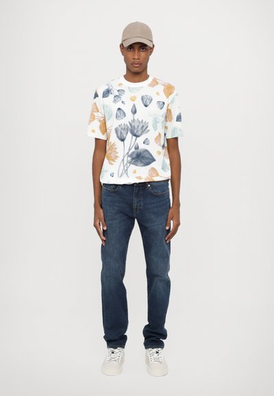 Short-sleeve white T-shirt with a floral pattern in blue, orange, and green, paired with dark blue jeans and white sneakers.