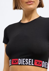 Svart avkortet t-skjorte laget av glatt stoff, med korte ermer og rund halsutskjæring, med et dristig rødt og hvitt logo-bånd nederst.