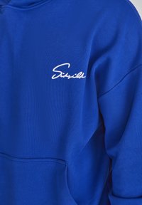Felpa blu con tasca a marsupio frontale, polsini a coste e logo bianco in corsivo sulla parte sinistra del petto. Tessuto morbido e texturizzato.