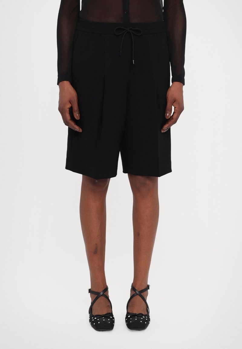 Černé šortky na míru s šňůrkovým pasem, plisovaným předním dílem a bočními kapsami, doplněné balerínami s puntíky. Hladká textura látky.