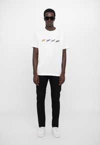 Biała bawełniana koszulka T-shirt z kolorowymi grafikami ryb na klatce piersiowej, w połączeniu z czarnymi spodniami i białymi trampkami. Model nosi okulary przeciwsłoneczne.