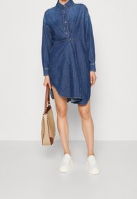 Denim hemdjurk met kraag, lange mouwen en knoopsluiting. Voorzien van een zijsplit en gecombineerd met een beige handtas met patroon en witte sneakers.