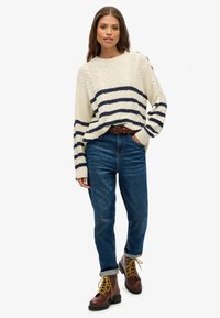 Strickpullover in Creme mit dunkelblauen Streifen und strukturiertem Muster; kombiniert mit blauen Jeans und braunen Schnürstiefeln. Verfügt über umgeschlagene Bündchen und einen Gürtel.