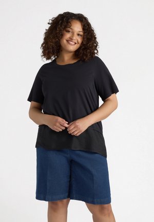 Camisa negra de manga corta con escote redondeado y un dobladillo ligeramente más largo. Combinada con pantalones cortos de mezclilla azul de longitud media. Textura de tela suave.
