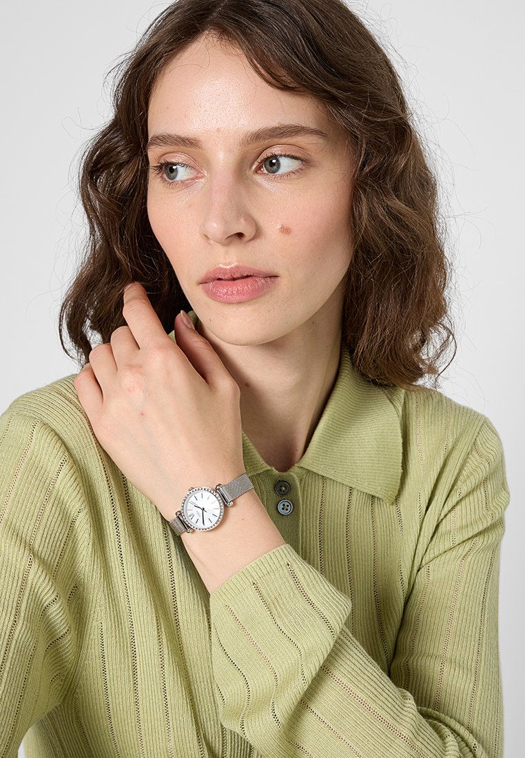 Orologio da polso in rete argentata con quadrante bianco, indicatori delle ore neri e lunetta decorata, indossato su una mano contro una camicia a coste verde chiaro.